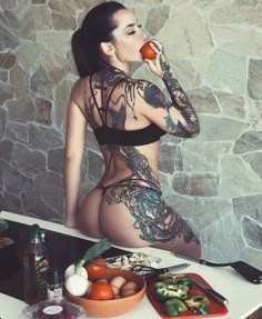 a woman with tattoos on her body sitting at a table eating an apple and vegetables