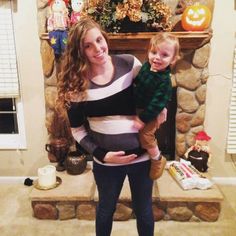 a woman holding a baby while standing in front of a fireplace