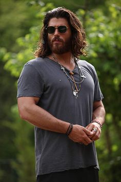 a man with long hair wearing sunglasses and a chain around his neck standing in front of trees