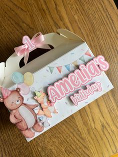 an opened birthday box on a table with a teddy bear sticker and pink ribbon