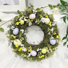 an easter wreath with eggs and flowers on a white table cloth next to greenery