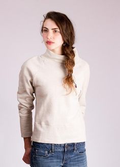 a woman with long hair wearing a white turtle neck sweater and jeans, standing in front of a gray background