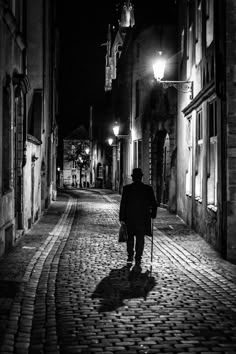 a man walking down a street at night with a cane in his hand and wearing a hat