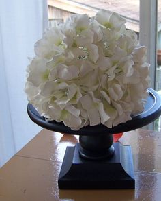 a black vase with white flowers in it on a wooden table next to a window