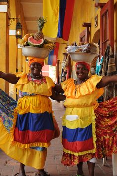 two women in brightly colored dresses and hats are posing for the camera with fruit on their heads