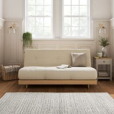 a white couch sitting in front of two windows next to a wooden table and rug