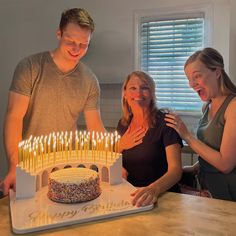 two women and a man standing in front of a birthday cake with candles on it