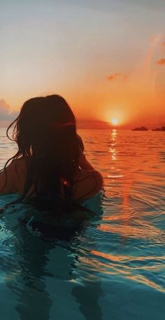 a woman in the water at sunset with her back to the camera, looking out into the distance