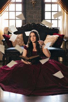 a woman in a long red dress sitting on a couch with papers flying around her