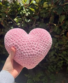 a hand holding a pink crocheted heart in front of some bushes and trees