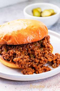 a sloppy joe sandwich on a white plate with pickles in the bowl behind it