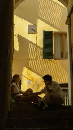 a man and woman sitting on the steps in front of a building with green shutters