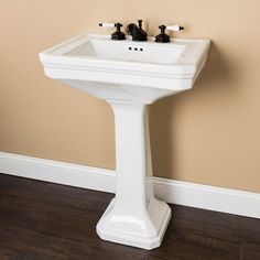 a white pedestal sink with two faucets on the side and a brown wall behind it