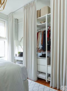 a white closet with curtains and clothes hanging on the door, next to a bed