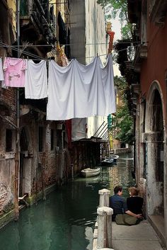 two people sitting on the edge of a narrow canal with laundry hanging over it and buildings in the background