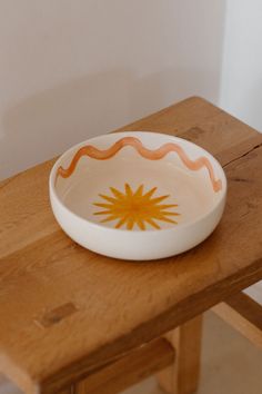 a bowl sitting on top of a wooden table next to a white wall with a yellow sun painted on it