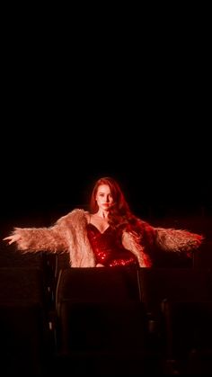 a woman sitting in a chair with her arms spread out, wearing a red dress and fur coat