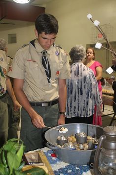 a man in uniform preparing food on a table with other people standing around and looking at it