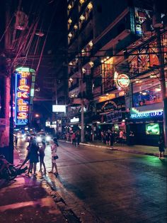 people are walking down the street at night with neon signs on the buildings in the background