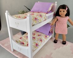 a doll standing next to a white bunk bed with pink and purple pillows on it