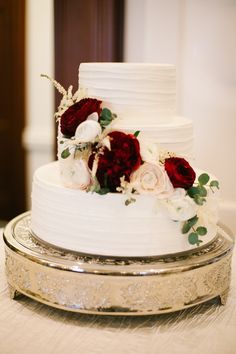 a white wedding cake with red and white flowers on the top tier is sitting on a silver platter