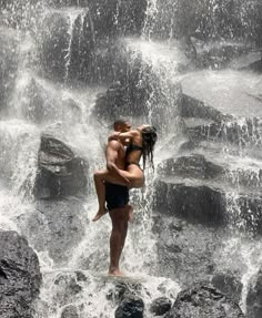 a man and woman are standing in front of a waterfall, one is holding the other