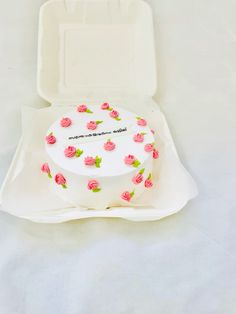 a white cake with pink roses on it sitting in a foam box next to a fork and knife