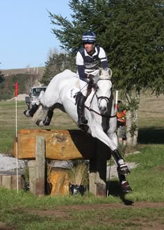 a man riding on the back of a white horse jumping over a wooden obstacle in a field