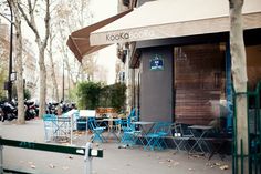 an outdoor cafe with blue chairs and tables on the sidewalk in front of it, surrounded by trees