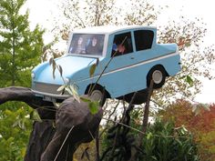 a blue car on top of a tree trunk with people in the window and behind it is an elephant statue