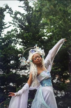 a woman dressed in white and blue is holding her arms out to the side with trees behind her