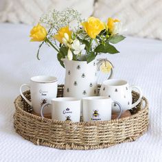 coffee cups and mugs are arranged in a wicker tray on a bed with yellow roses