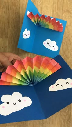 someone is holding an origami fan with clouds on it and rainbows in the sky