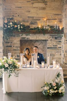 a bride and groom sitting at a table in front of a fireplace with candles on it