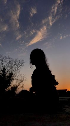 the silhouette of a person sitting in front of a tree at sunset with clouds overhead