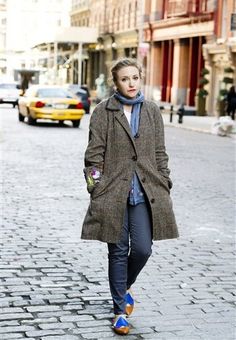 a woman is walking down the street wearing a coat and blue shirt with orange shoes