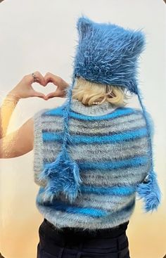 a woman making a heart with her hands while wearing a fuzzy blue hat and sweater