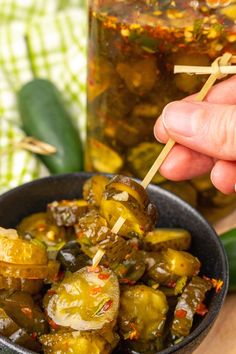 a hand holding a toothpick over some pickles in a bowl next to a jar