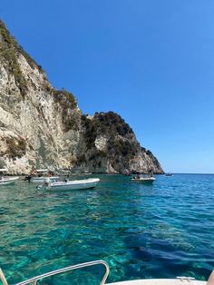 several small boats floating in the water near a rocky cliff face and clear blue ocean