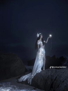 a woman in white dress standing on rocks at night holding a light up item with her right hand