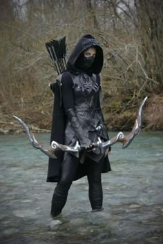 a woman dressed in black and holding two arrows, standing in the water with trees behind her