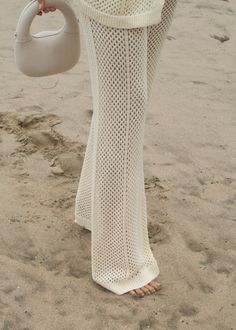 a woman walking on the beach carrying a white purse and wearing a long knitted dress