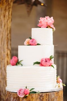 a three tiered cake with pink flowers on top is sitting on a tree stump