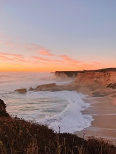 the sun is setting over the ocean with waves crashing on the shore and cliffs in the background