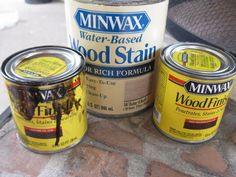 three cans of wood stain sitting next to each other on a brick patio area with a metal grill in the background