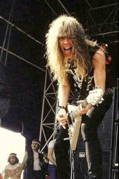 a man with long hair playing guitar on stage while other people watch from the sidelines