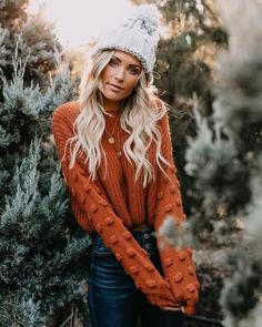 a woman standing in front of some trees wearing a hat and sweater with her hands on her hips