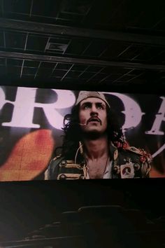 a man with long hair standing in front of a large screen displaying the movie's character