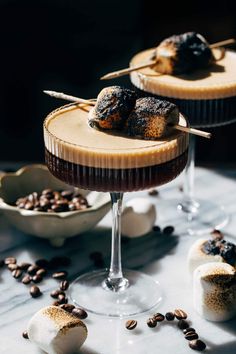 two desserts with chocolate and marshmallow toppings on a table next to coffee beans