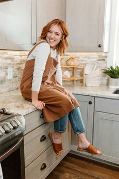 a woman sitting on the counter in a kitchen smiling at the camera while wearing an apron